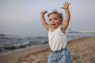 A baby in summery clothes and flowers in her hair stands on the beach at sunset and stretches both