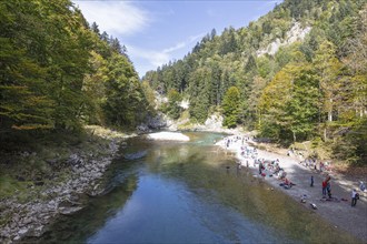 A river beach with people sitting by the water, surrounded by autumnal trees and forests,