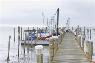 A wooden dock extends out into the water with fishing boats moored alongside under a misty, cloudy