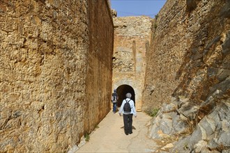 Two people walking through a narrow passageway between high stone walls of an ancient fortress,