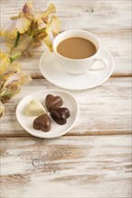 Cup of cioffee with chocolate candies, lilac and purple iris flowers on white wooden background.