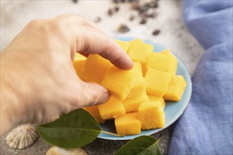 Dried and candied mango cubes on blue plate with hand on brown concrete background and linen