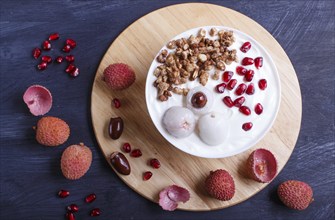 Greek yogurt with lychee, pomegranate seeds and granola in a white plate on a black wooden