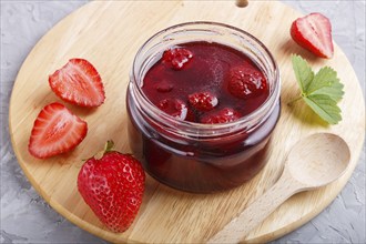 Strawberry jam in a glass jar with berries and leaves on gray concrete background. Homemade, close