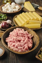 A wooden bowl filled with raw ground meat is surrounded by pasta, mushrooms, garlic, and cheese on