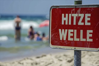 Warning sign with German text 'Hitzewelle' (Heat wave) in front of blurry sunny beach with people.