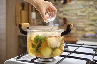 A hand adds vodka to a pot with fish soup, vegetables and herbs on a stove. The kitchen features a