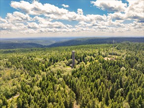 An observation tower stands in the centre of a vast green forest under a cloudy blue sky,