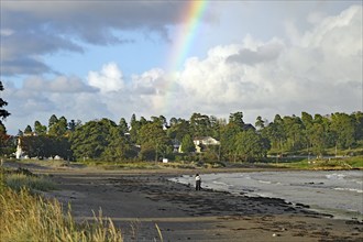 Beach landscape with rainbow and clouds over the sea and scattered green trees in the background,