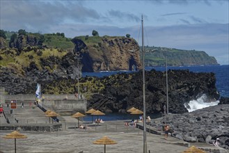 A rocky coastline with people in a seawater swimming pool resting under parasols while the sea