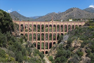 An old brick aqueduct spans a wooded ravine against a mountain backdrop under a clear blue sky,