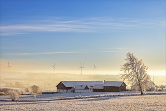 Farmhouse in a field with snow and frost and low cold fog in a valley and wind turbines on the