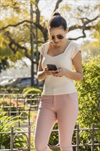 A young woman wearing sunglasses and casual dress looks at her smartphone while strolling outdoors