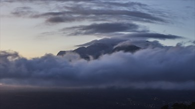 Mystical scene of a mountain peak shrouded in clouds at dusk with lights of a town in the valley,