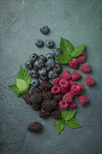 A mix of ripe berries, with leaves scattered on the table, raspberries, blackberries, blueberries,