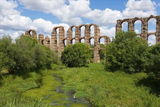 Antike Aquädukt-Ruinen inmitten von üppiger Vegetation und Fluss unter blauem Himmel, Acueducto de