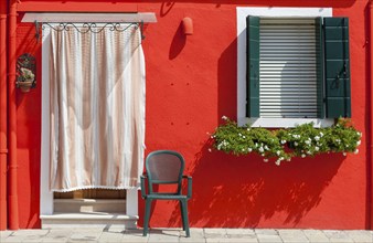 Red house wall with shutters, curtain, green chair and flower box, Burano, Venice, Italy, Europe