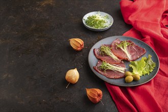 Slices of smoked salted meat with cilantro microgreen on black concrete background and red textile.