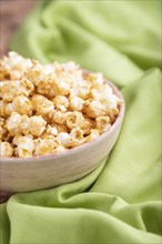 Popcorn with caramel in ceramic bowl on brown concrete background and green textile. Side view,