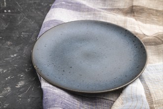 Empty blue ceramic plate on black concrete background and linen textile. Side view, close up