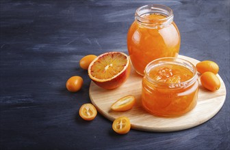 Orange and kumquat jam in a glass jar with fresh fruits on black wooden background. Homemade, close