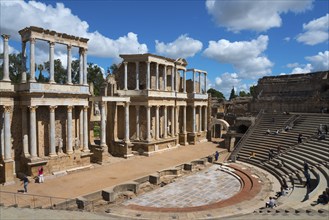 Wide view of a Roman theatre with ancient ruins and spectators on the steps under a blue sky,