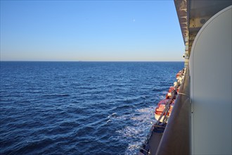 A large ship sails calmly on the blue ocean under a clear sky, with a view of the lifeboats from