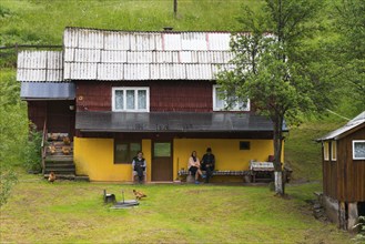 A yellow house surrounded by green grass with relaxed people and chickens in the front yard, Old