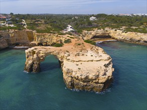 Large, rocky coastline with a natural arch surrounded by clear water and green vegetation, aerial