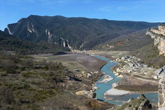 A winding river through a wide valley, surrounded by wooded mountains and rocks under a clear sky,