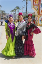 Three woman in colourful flamenco dresses, with flowers in their hair, pose on a sunny street with