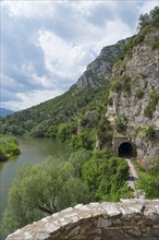 A river winds along a mountain with a railway tunnel, surrounded by lush vegetation under a cloudy