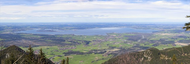 Panoramic view over a green landscape with mountains in the foreground and the large Chiemsee in