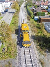 Yellow vehicle on rails in a residential area under partly sunny, partly cloudy skies, tamping