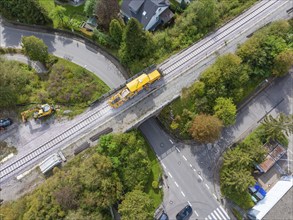 A yellow construction machine on railway tracks crosses a bridge on a winding road in a green