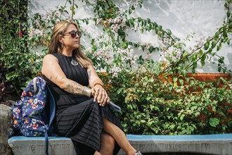 A fashionable woman with tattoos and a flower backpack sits on a bench, surrounded by vibrant