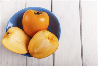 Ripe orange persimmon in a blue plate on white wooden background, with copy space