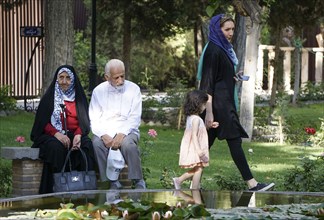 A young Iranian woman walks with her daughter past a traditionally dressed elderly couple sitting