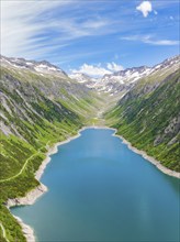 Landscape with a long, blue lake surrounded by high grass and tree-covered hills and snow-capped