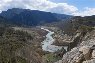 A river meanders through a wide valley surrounded by green hills and rocky mountains under a partly