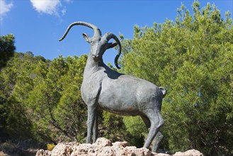 Bronze statue of an ibex in nature on a rock base under a blue sky, Área Recreativa de Sedella,