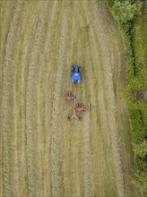 Aerial view of a tractor with a hay tedder working a hay field, surrounded by trees and a country