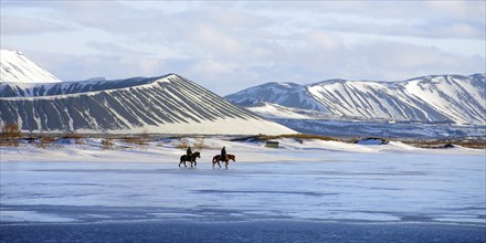 Two riders on horses on a partly frozen ice surface in a snow-covered winter landscape, volcanoes