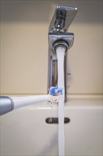 A bathroom tap is running while a toothbrush is held under the running water, symbolic image of