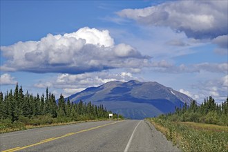 An empty road leads through a picturesque landscape with mountains and clouds in the blue sky,