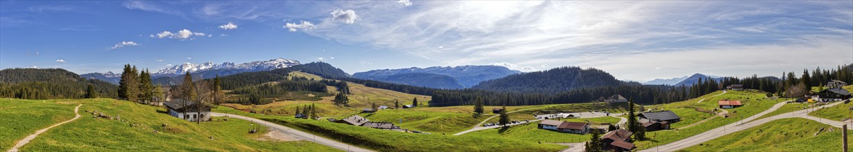 Wide green meadows in front of a mountain range, dotted with huts, under a partly cloudy blue sky,