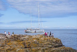 People on rocks by the sea with sailboat in the background in Gudhjem, Bornholm, Baltic Sea,