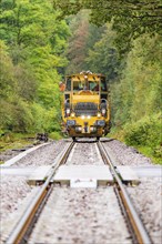 The yellow tractor unit approaches on a railway track, flanked by green forest, Hermann Hesse Bahn