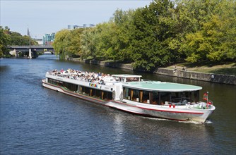 A large excursion boat sails on a river through a green, urban environment in sunny weather,