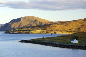 A single house stands on the shore of a tranquil inlet, surrounded by mountains and a blue sky,